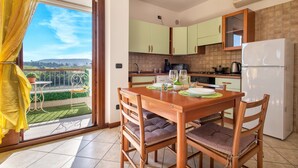 Ein Essbereich mit Tisch und Stühlen, Blick ins Freie, eine Küche mit Schränken und Geräten sowie ein Balkon mit Geländer und Topfpflanzen.