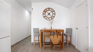A dining area with a wooden table, four chairs, a wall clock, and a small vase with flowers.