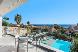 A balcony with a pool, chairs, and a table, overlooking a coastal area with houses and palm trees.