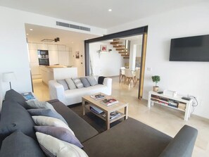 A modern living room with a sofa, coffee table, and a TV. There is a kitchen in the background and a dining area with chairs.