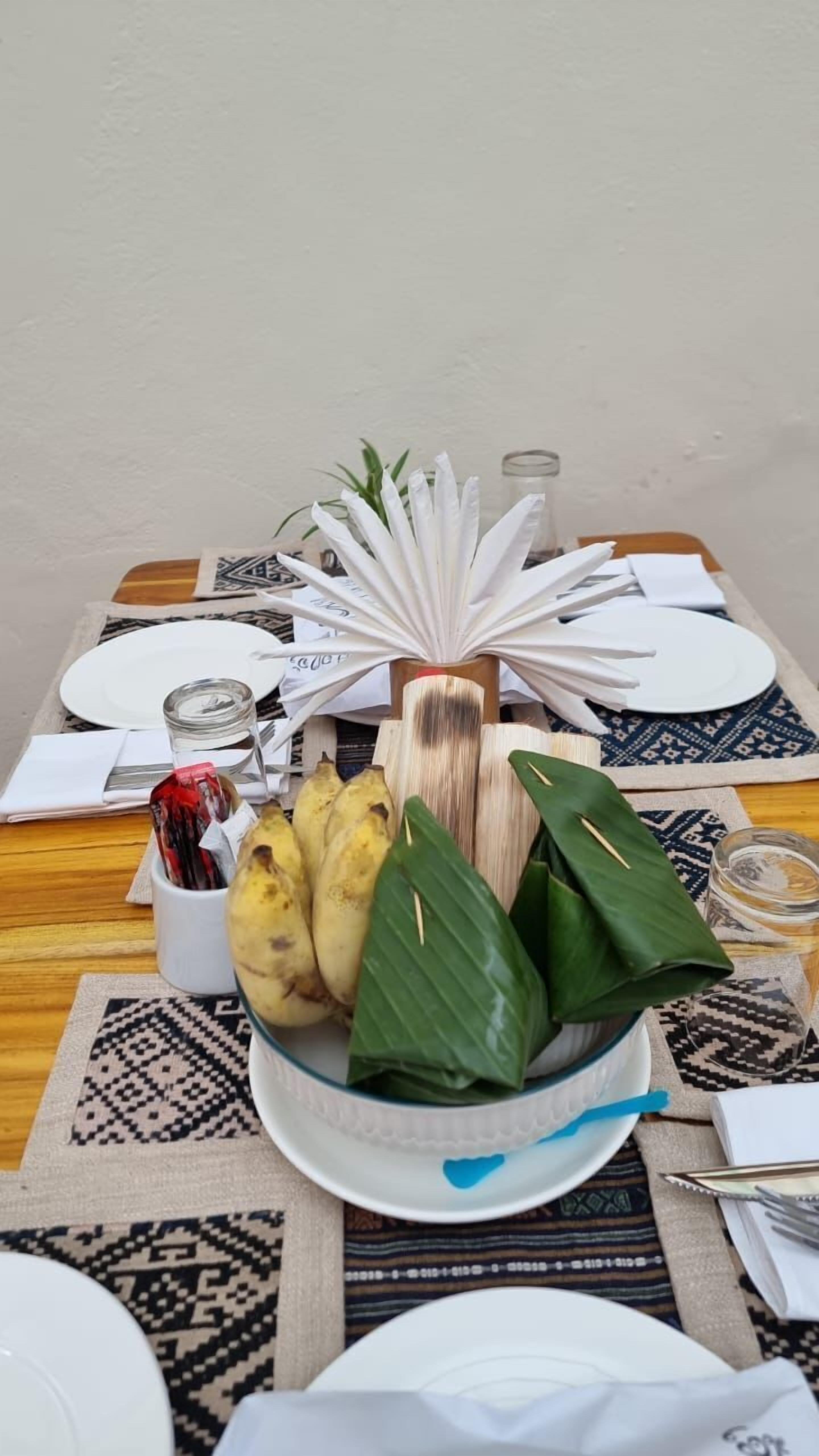 A table set for a meal with a central bowl containing bananas and a bottle, surrounded by plates and cutlery.