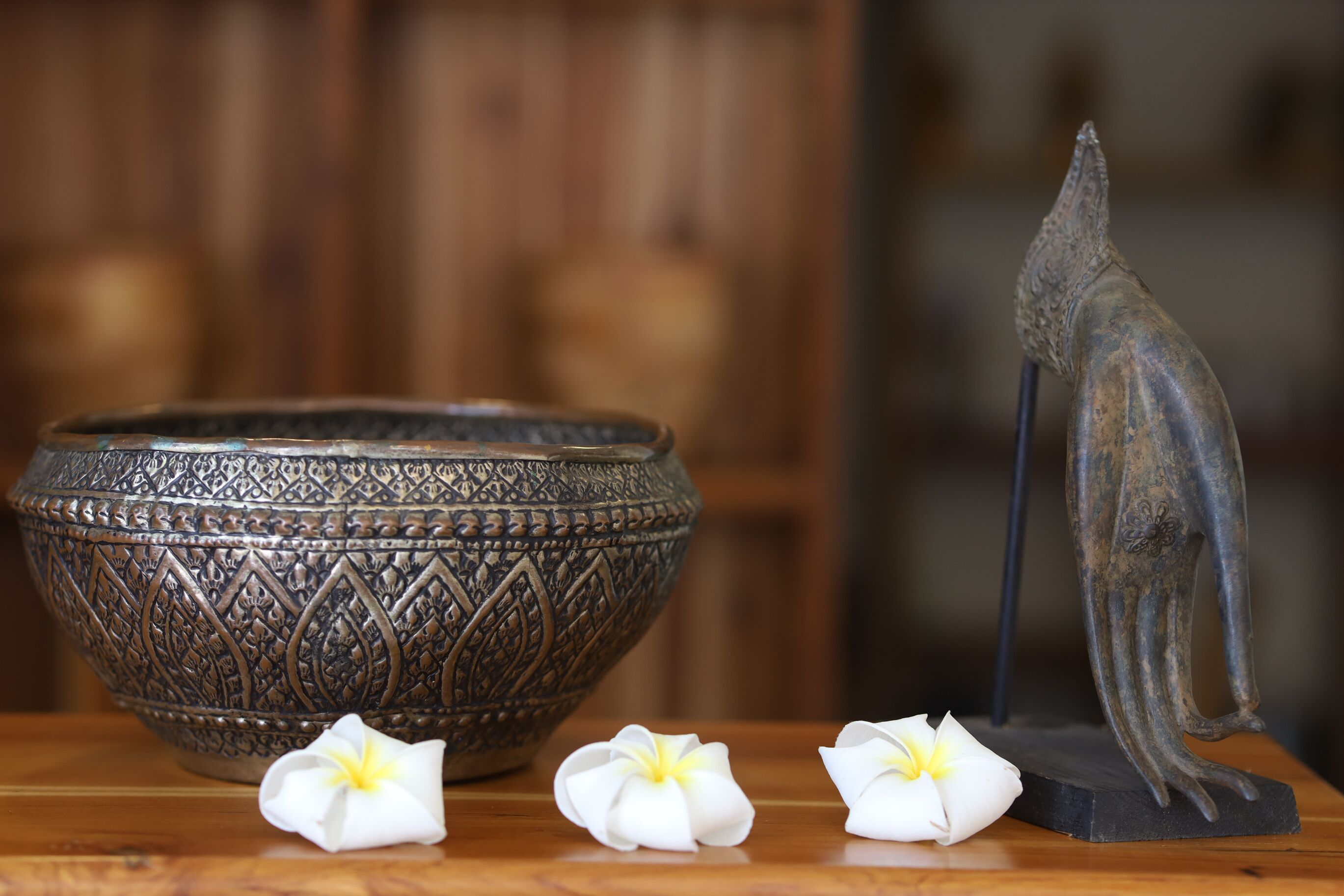 A decorative bowl with intricate patterns, a small statue, and two white flowers on a wooden surface.