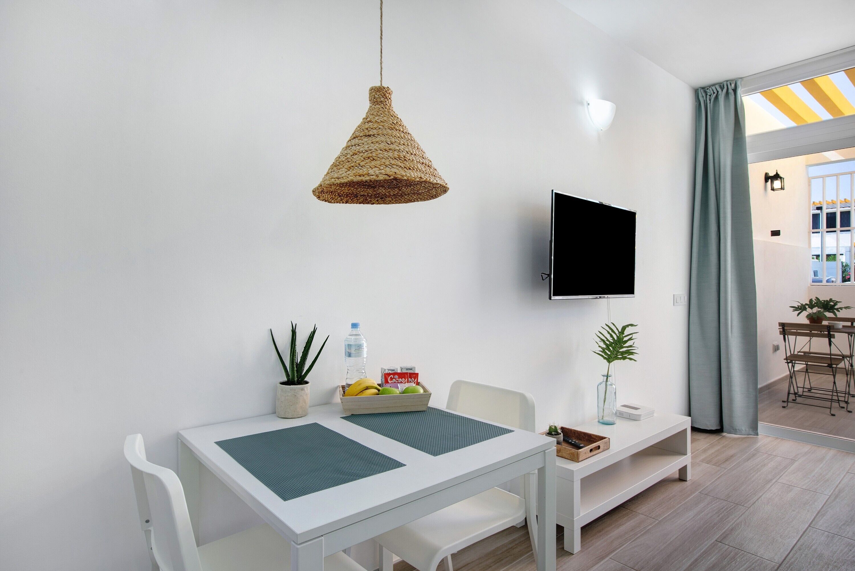 A modern room with a white table, a woven pendant light, a flat-screen TV, a potted plant, and a view of the outside.