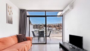 Uma sala de estar moderna com sofá, televisão de tela plana, mesa de jantar com cadeiras e uma porta de vidro deslizante que dá para um terraço ao ar livre.