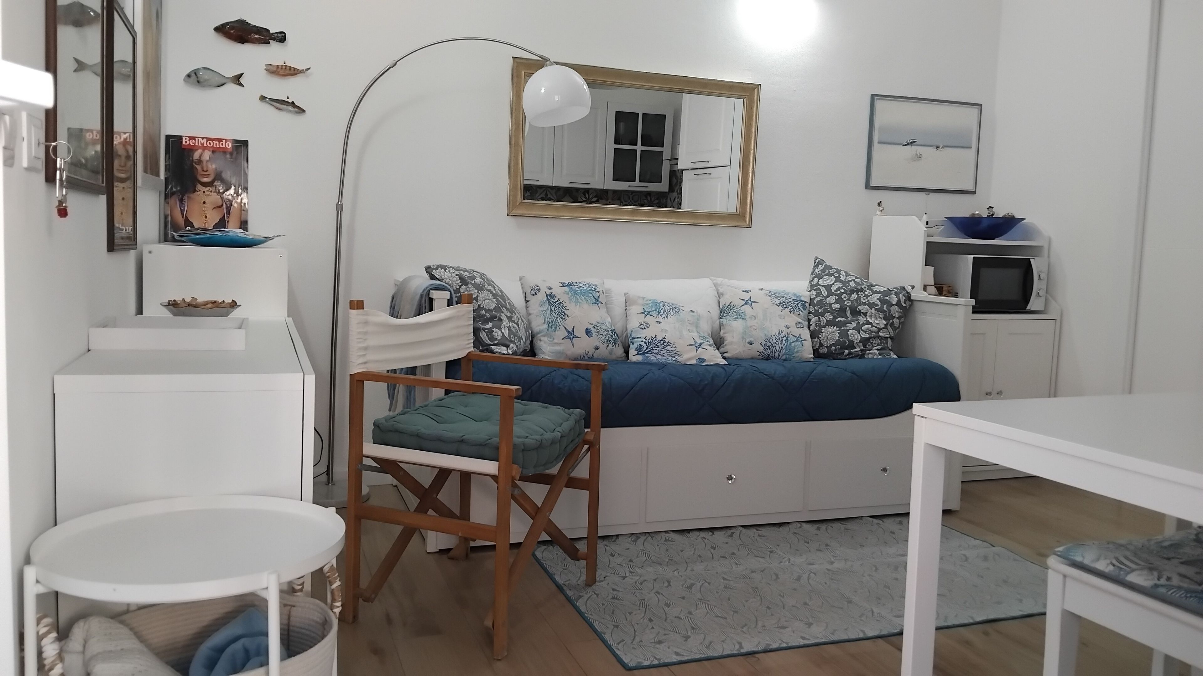 A compact bedroom with a bed, a desk, a chair, a mirror, and a small kitchenette.