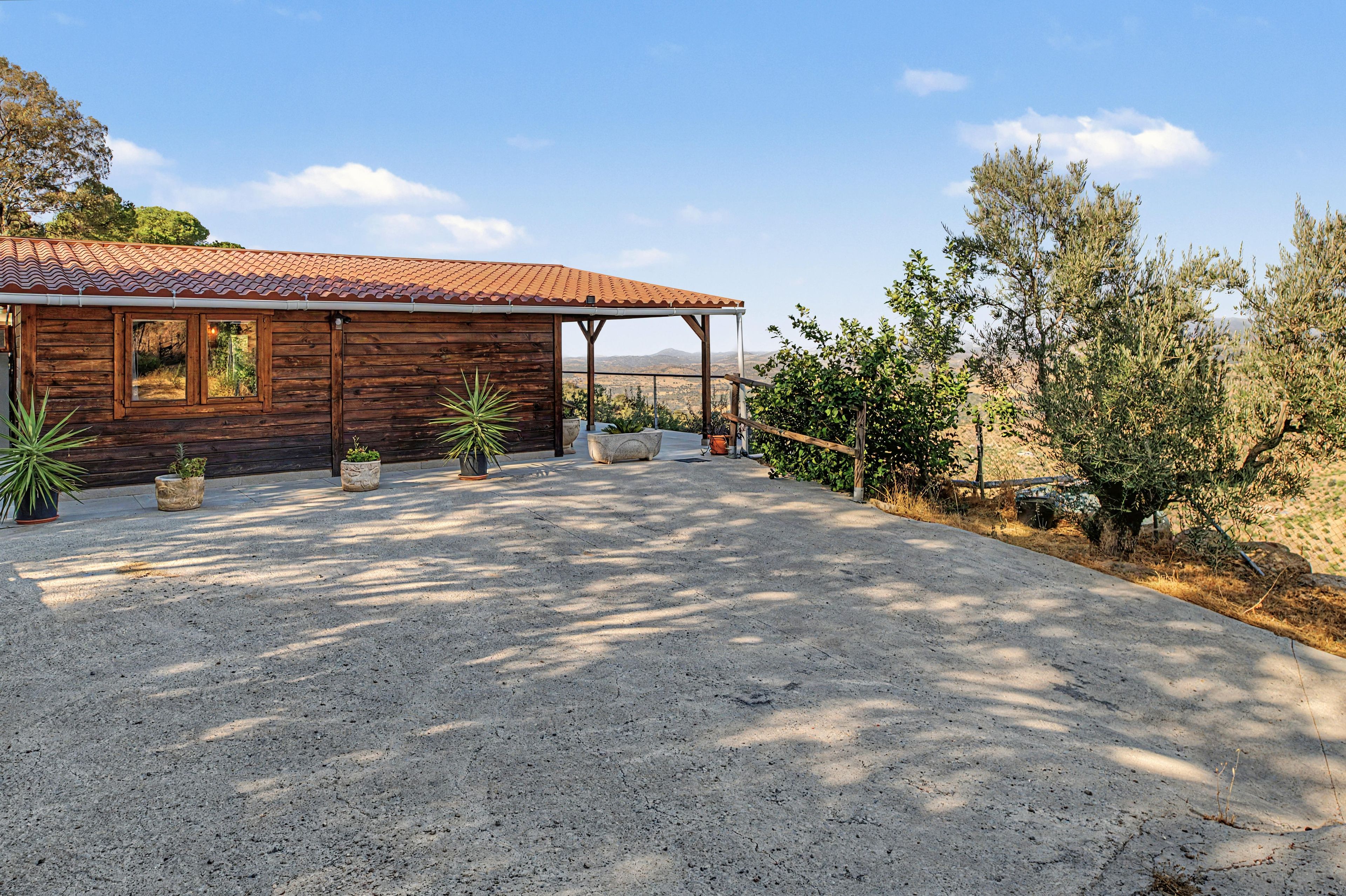 A wooden cabin with a tiled roof, surrounded by potted plants and a gravel driveway.
