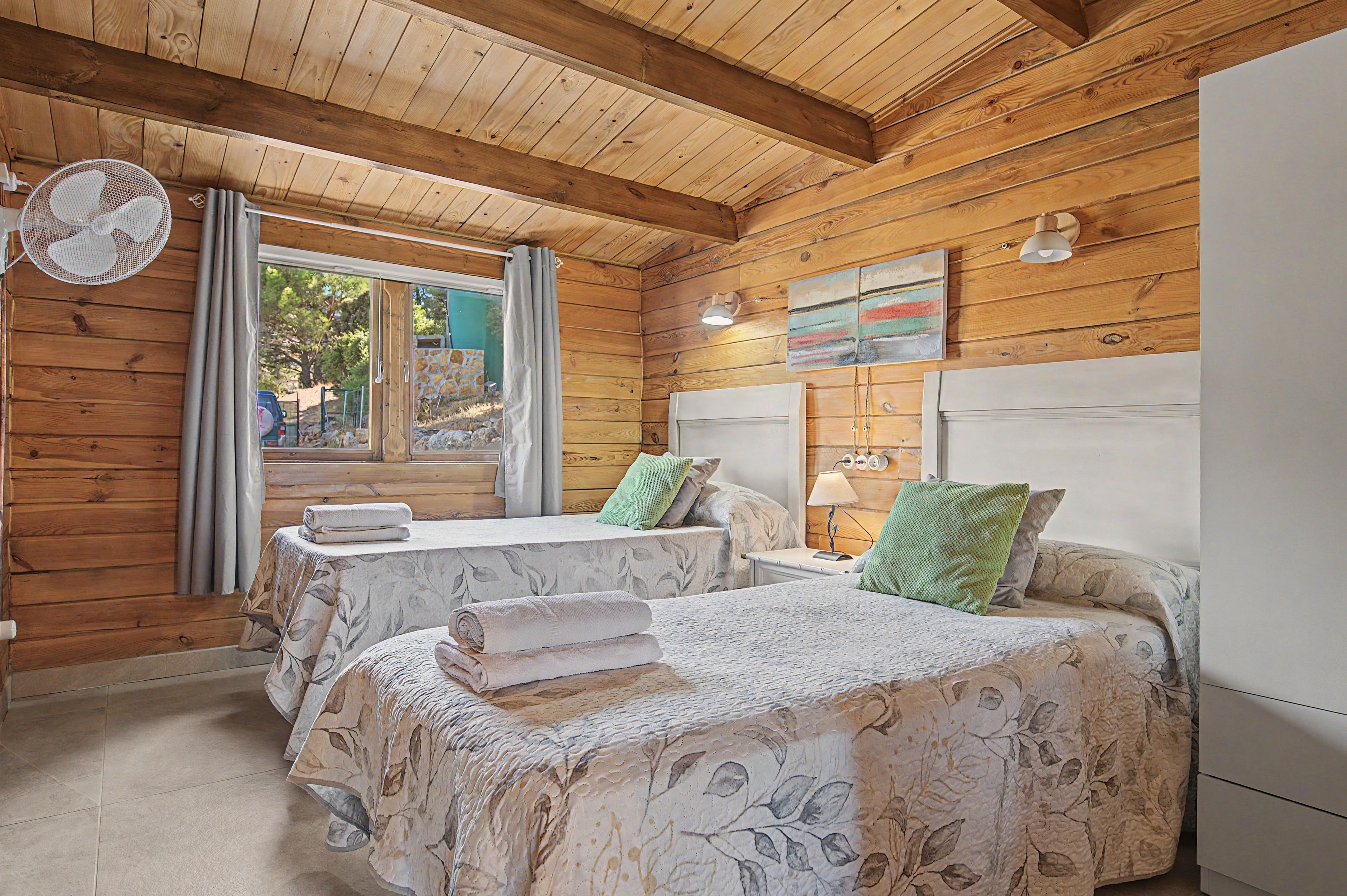 A wooden cabin room with two beds, a fan, and a window with curtains.