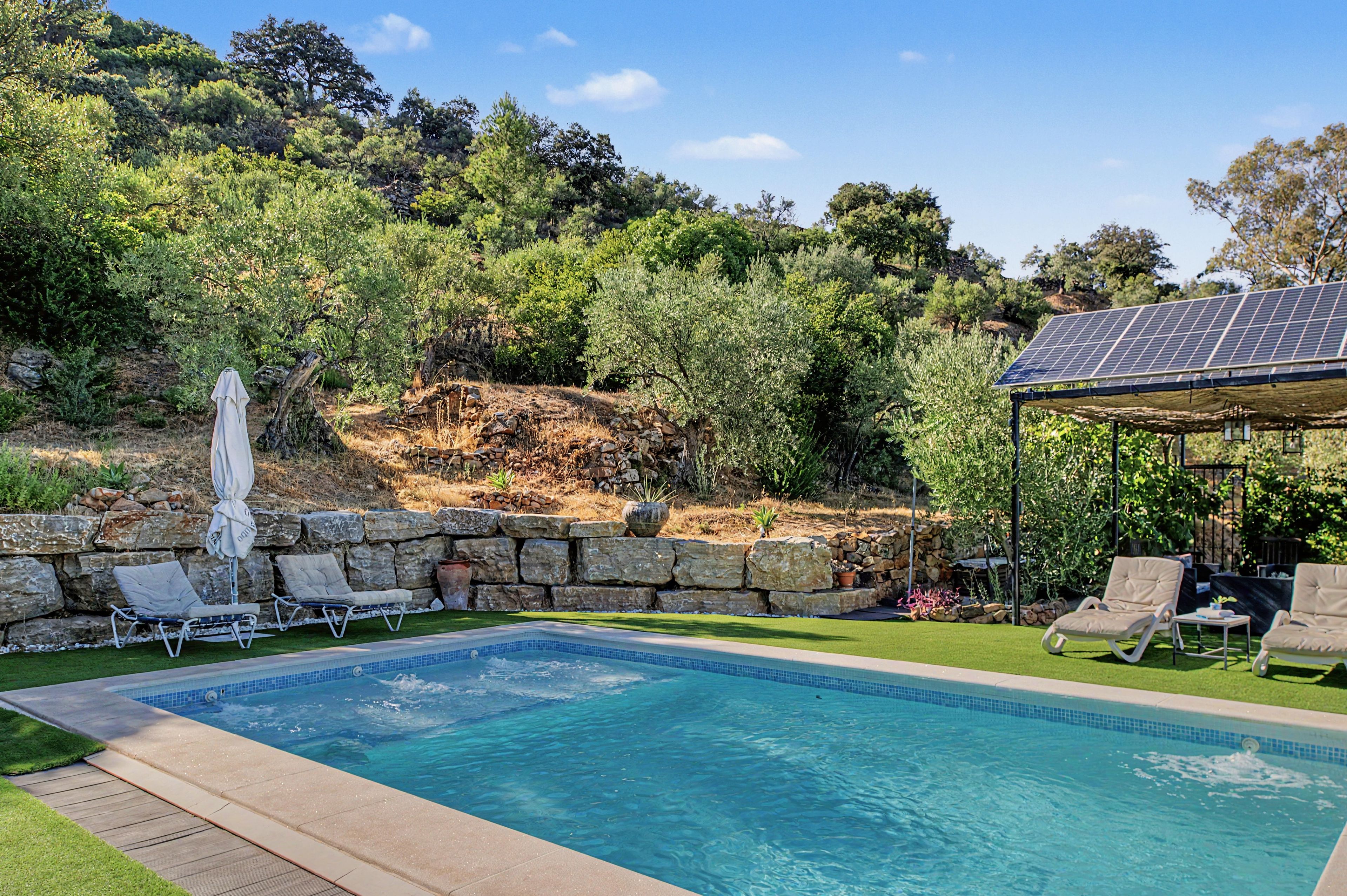 A swimming pool with lounge chairs, a stone wall, and a solar panel structure.