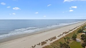 A beach with a long stretch of sand, a pier in the distance, and a clear sky.