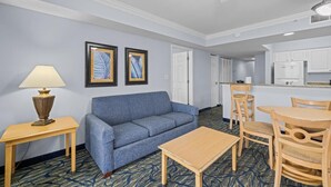A hotel room with a blue sofa, a wooden table, a lamp, and a kitchenette. - Sunset Retreat: Two Bdrm Oceanfront 15th Floor (Myrtle Beach)