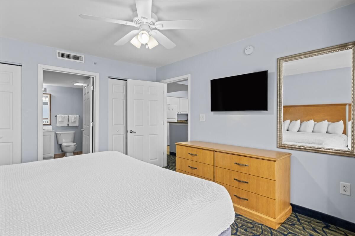 Une chambre d’hôtel avec un grand lit, une commode en bois, un ventilateur de plafond, un téléviseur fixé au mur et un miroir avec un cadre en bois.