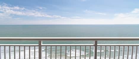 Une vue sur la plage depuis un balcon doté d’une balustrade, avec les vagues de l’océan et un ciel dégagé.