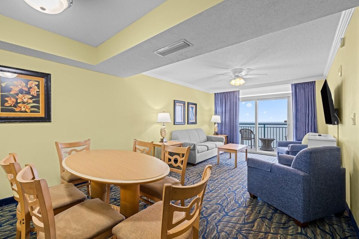 A hotel room with a dining table, chairs, a sofa, a loveseat, a TV, and a balcony with a view.
