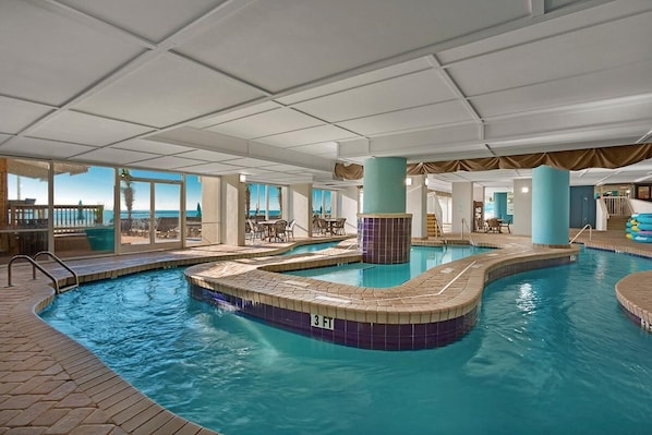 An indoor swimming pool with a 3 FT depth marker, surrounded by lounge chairs and a view of the ocean through large windows.