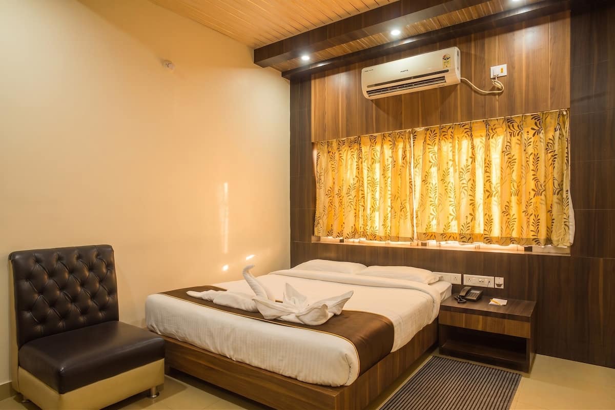 Una habitación de hotel con cama, una silla, equipo de aire acondicionado, ventana con cortinas y mesita de noche con teléfono y lámpara.