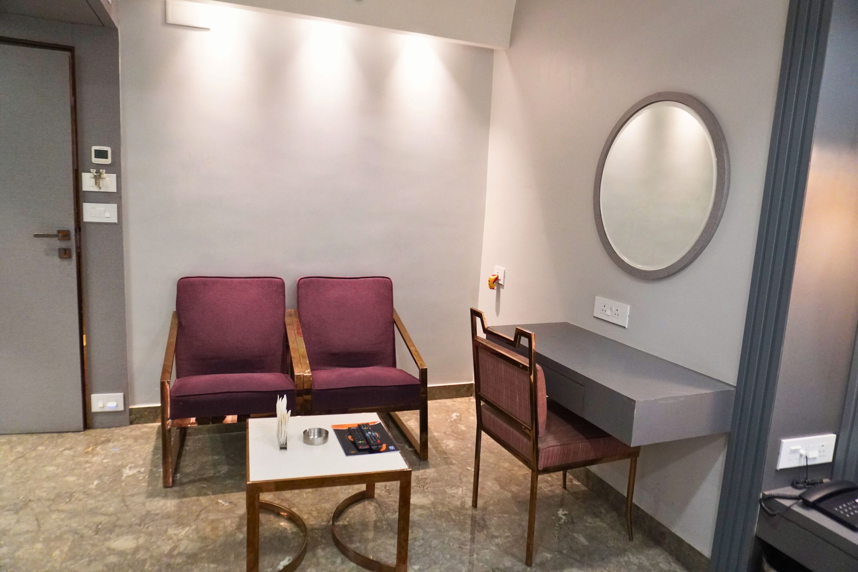 Quarto com duas cadeiras roxas, uma mesa pequena, escrivaninha, espelho e telefone em uma escrivaninha.