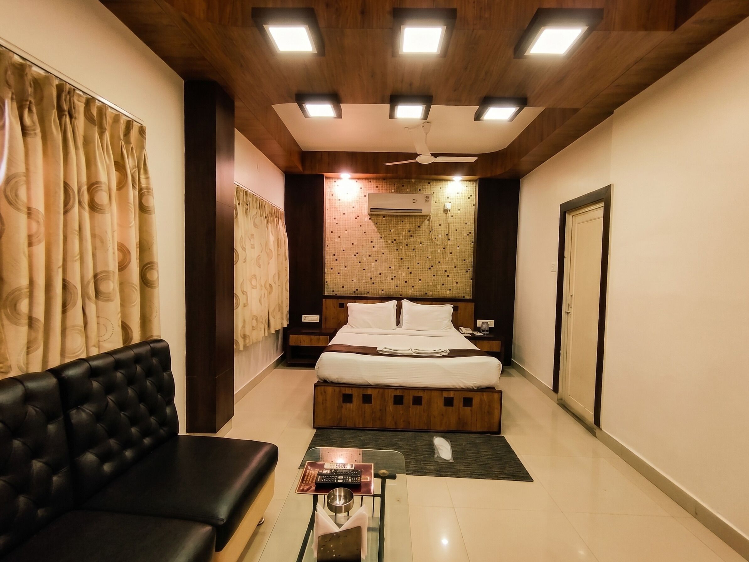 A hotel room with a bed, a black leather sofa, a small table with a remote control, and a wall-mounted air conditioner.