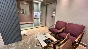 A modern hotel room with a marble floor, a small table with a remote control, two purple armchairs, a bench, and a kitchenette area. - continental lodge  (Malda)
