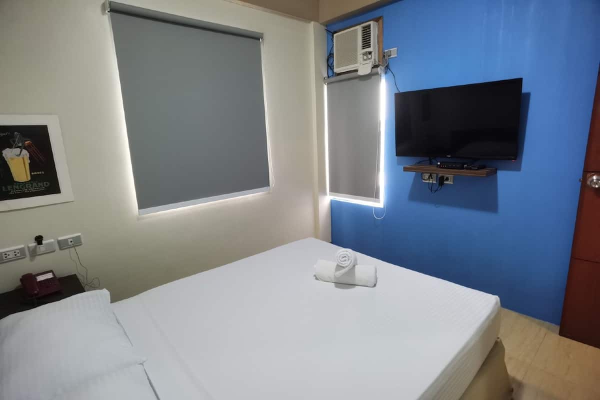 A hotel room with a bed, a TV mounted on the wall, a window with blinds, an air conditioning unit, and a telephone on the nightstand.