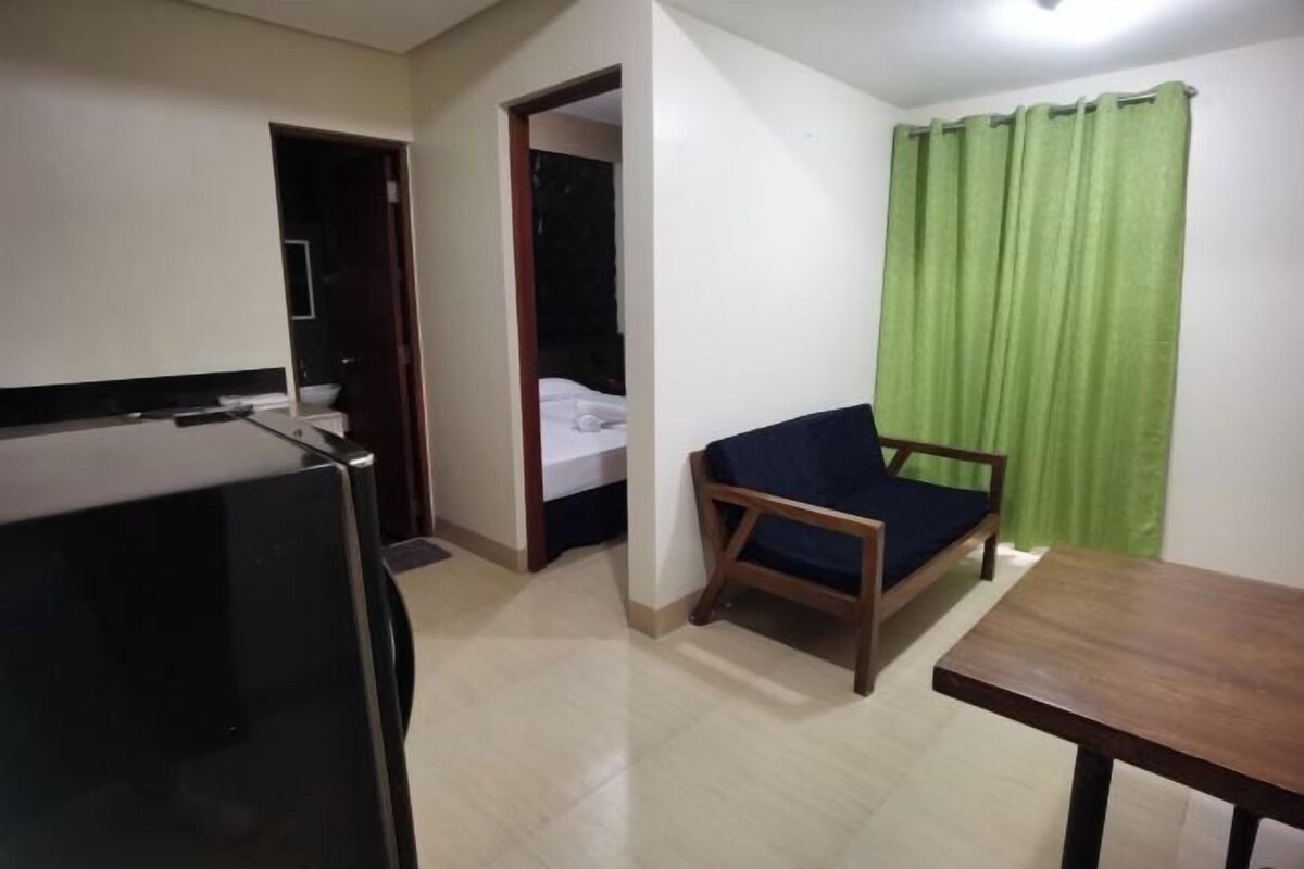 Una habitación pequeña con kitchenette, una cama individual, una silla de madera y una cortina verde.