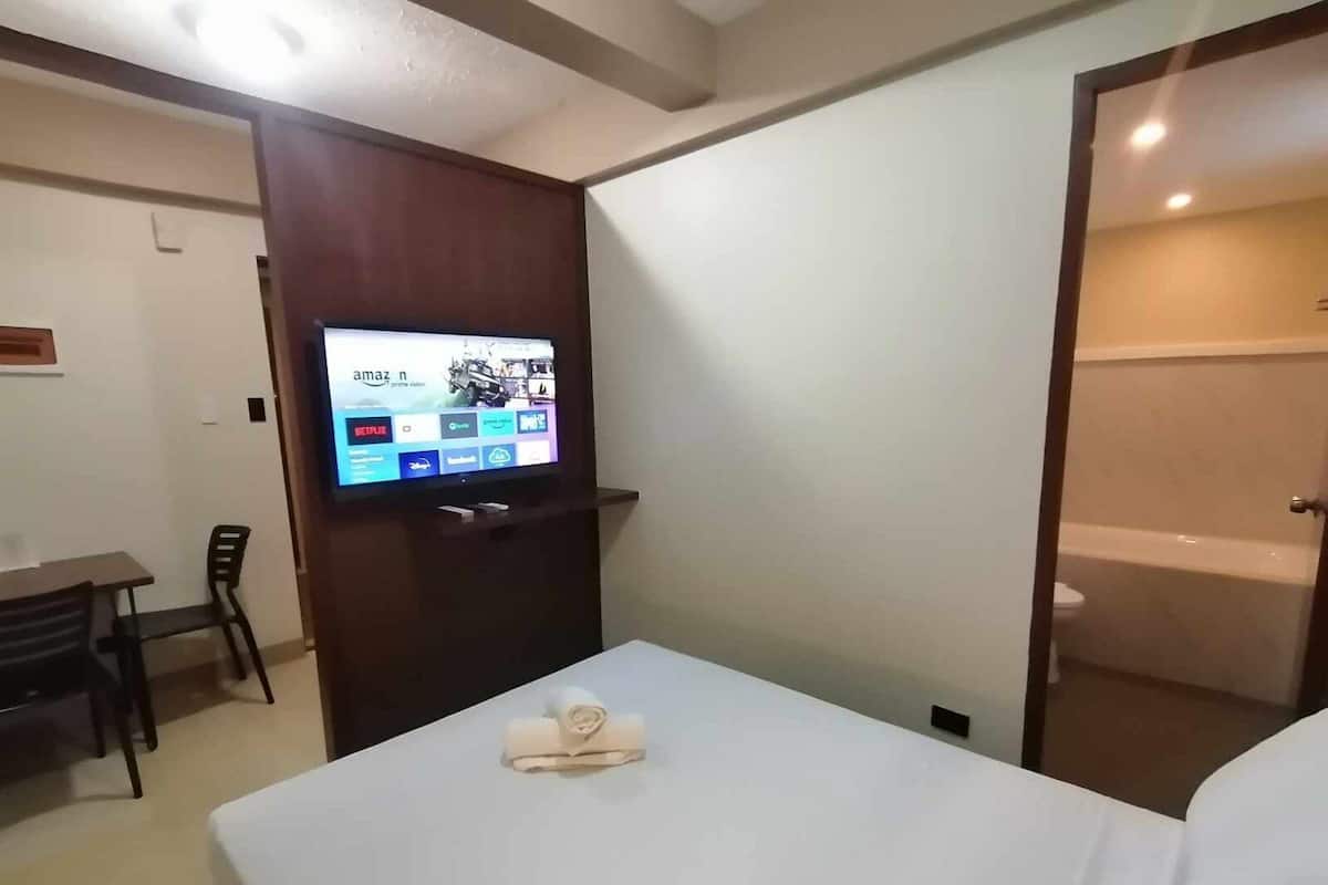 A hotel room with a bed, a TV mounted on a wall displaying various streaming services, a small table with chairs, and a bathroom visible through an open door.