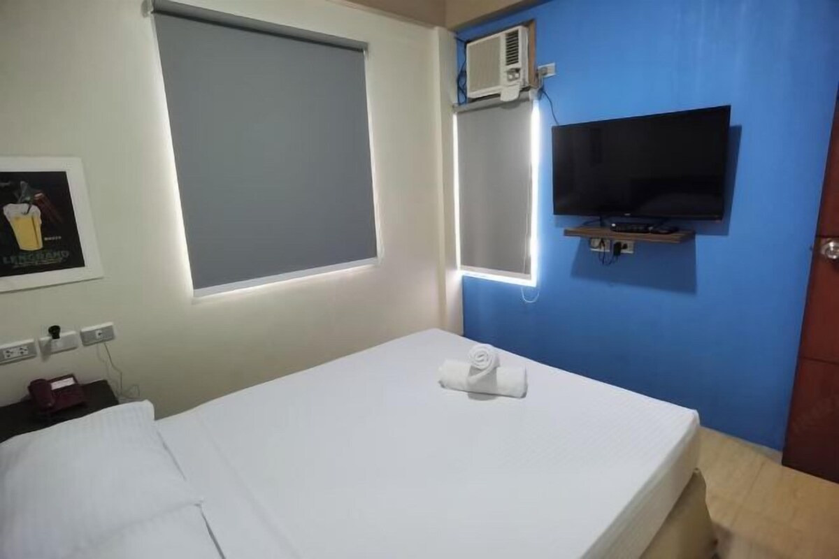 A hotel room with a bed, a TV mounted on the wall, an air conditioning unit, and a window with blinds.