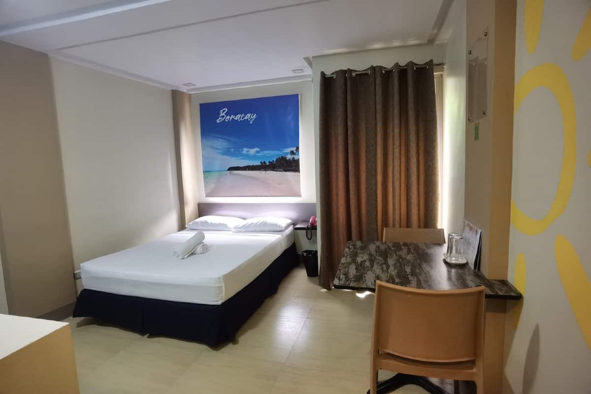 A hotel room with a bed, a small table with a chair, a window with curtains, and a wall with a beach poster.