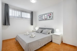 Un lit bien fait, avec des oreillers et une serviette pliée, une table de chevet avec une lampe, une fenêtre avec des rideaux et un mur orné de trois tableaux encadrés.