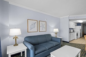 Un salon avec un canapé bleu, deux petites tables d’appoint blanches surmontées de lampes, et des œuvres d’art encadrées accrochées au mur. On aperçoit une cuisine au fond.
