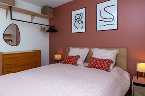 Une chambre avec une commode en bois, un lit agrémenté d’oreillers à motifs rouges et des œuvres d’art abstraites accrochées au mur.