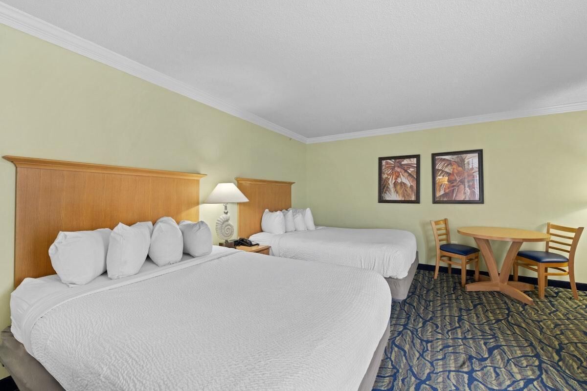 Une chambre d’hôtel avec deux lits, un bureau, des chaises, une lampe et deux tableaux encadrés au mur.