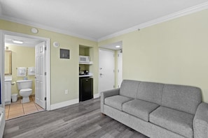 Un espace de vie compact comprenant un canapé gris, un coin cuisine avec un four à micro-ondes, et une salle de bain avec des toilettes et un lavabo.