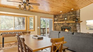 A cozy living room with a stone fireplace, wooden ceiling, and a dining area with a foosball table. - * Snowy Evening Lodge * / Treetop Views & Firepit (Big Bear)