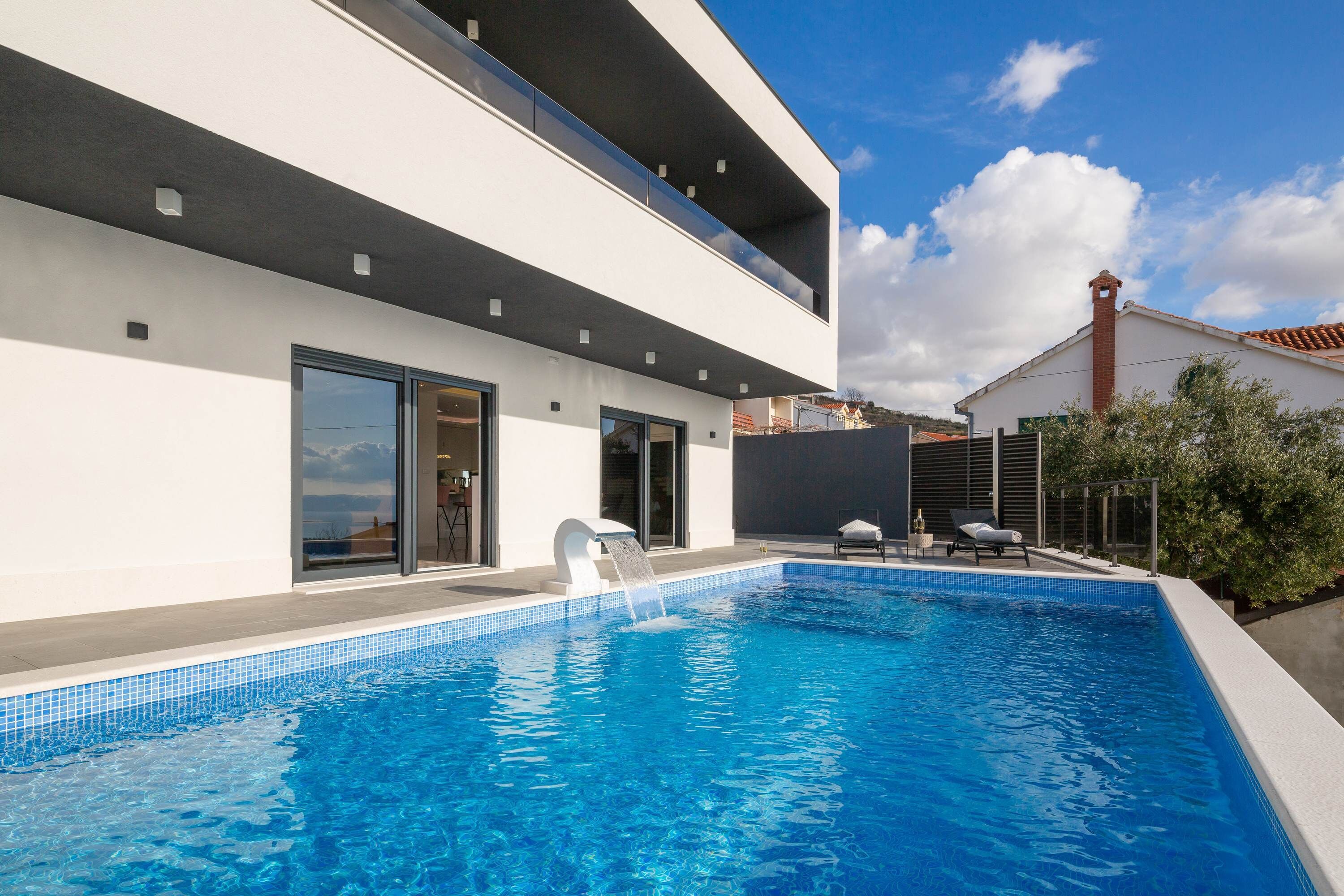 Un área de piscina moderna con una piscina azul cristalina, una fuente de agua y un edificio blanco con amplias ventanas y un balcón.