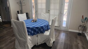 Un coin repas avec une table recouverte d’une nappe à carreaux bleus et blancs, entourée de chaises blanches. On y trouve deux portes avec des stores blancs, un présentoir métallique et une petite statuette de coq décorative.