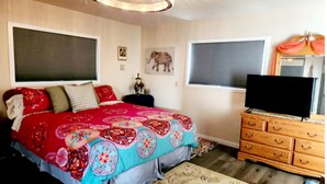 Une chambre à coucher avec un grand lit, une commode en bois, un téléviseur et une fenêtre avec des stores.