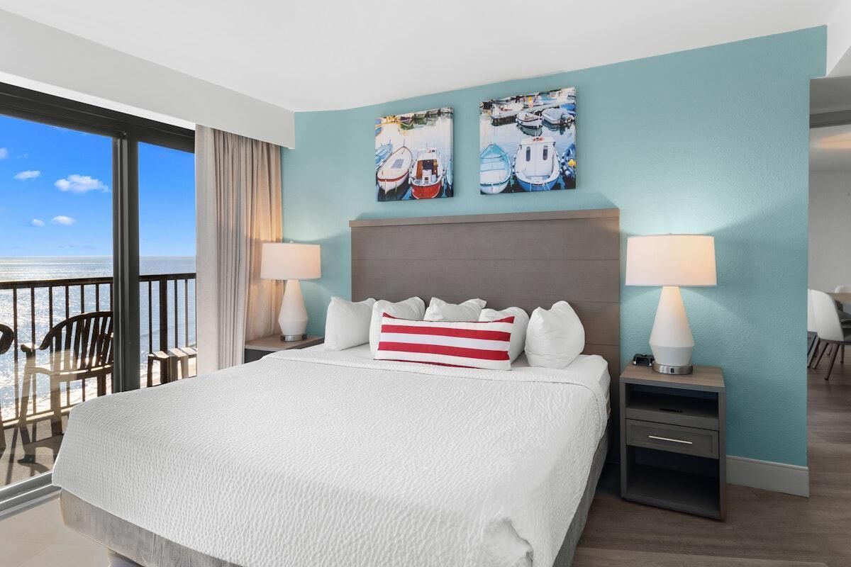 A hotel room with a large bed, two bedside lamps, a view of the sea, and two artworks on the wall.