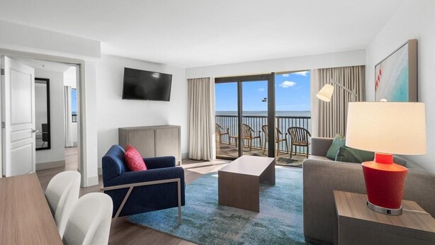 A modern hotel room with a balcony, a flat-screen TV, a sofa, a coffee table, and a lamp.
