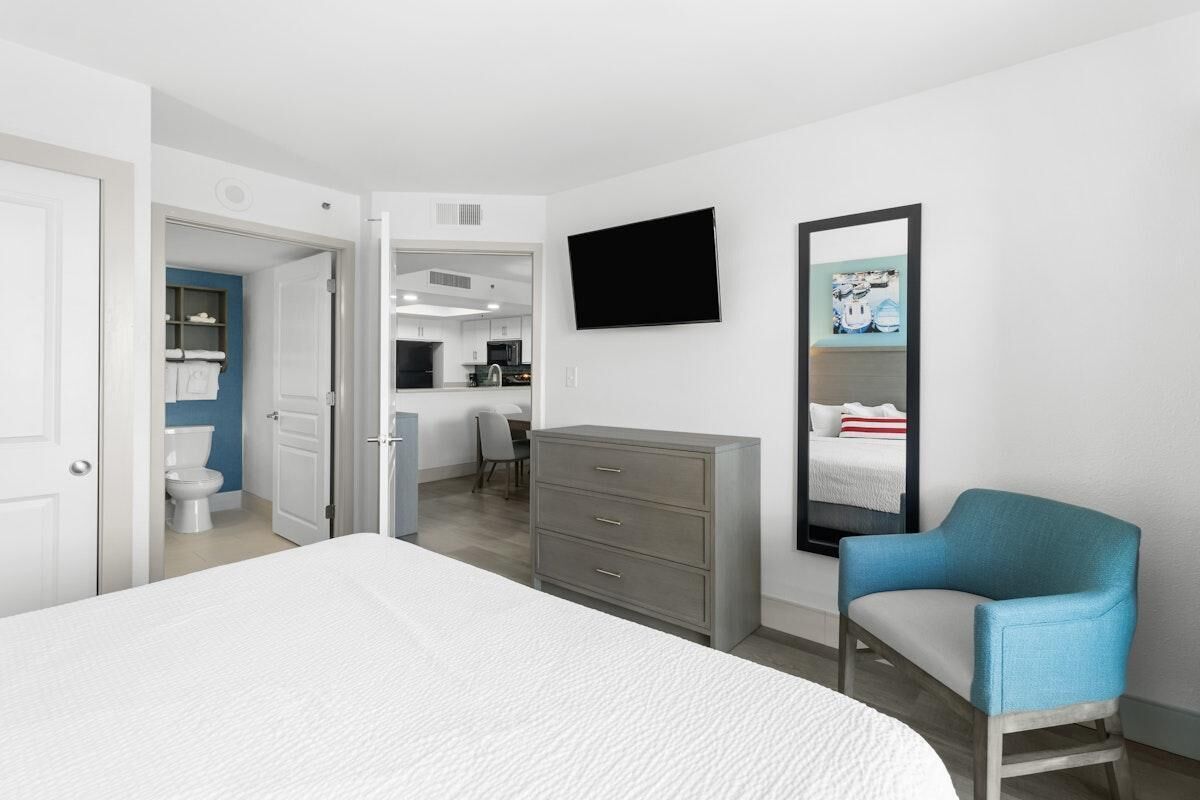 A modern hotel room with a large bed, a grey dresser, a blue armchair, a flat-screen TV, and a bathroom visible through an open door.