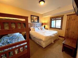 A bedroom with a large bed, a smaller bed with a patterned bedspread, a wooden headboard, a window with blinds, and a ceiling light.
