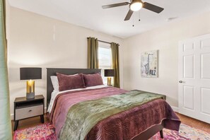 A bedroom with a bed, nightstand, lamp, ceiling fan, window with curtains, and a door.
