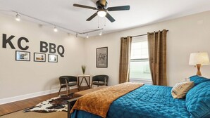 A bedroom with a bed, a ceiling fan, a desk with chairs, a cowhide rug, and a wall with 'KC BBQ' signage. - The Pioneer - Charming Home Walkable to Streetcar! (Kansas City)