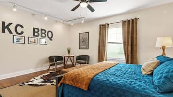 Une chambre avec un lit, un ventilateur de plafond, un bureau avec des chaises, un tapis en peau de vache et un mur orné d’une enseigne indiquant « KC BBQ ».