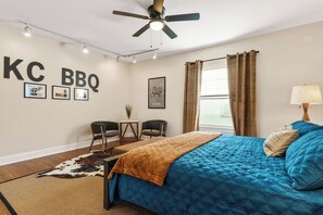 Un dormitorio con una cama, un ventilador de techo, un escritorio con sillas, un tapete de cuero de vaca y una pared con letrero 'KC BBQ'.