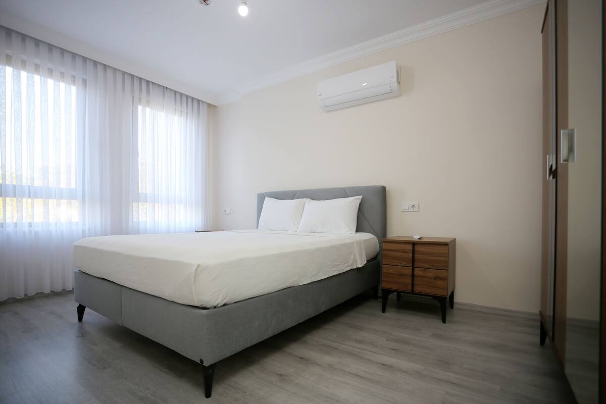 Ein modernes Schlafzimmer mit einem grauen Bett, weißen Bettwäsche, einem Holznachttisch und einer Klimaanlage an der Wand.