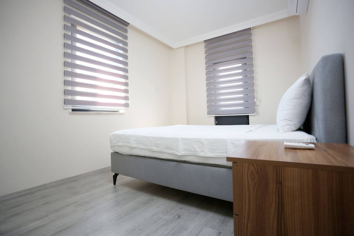 Ein Schlafzimmer mit einem Bett, einem Nachttisch aus Holz und zwei Fenstern mit Jalousien.