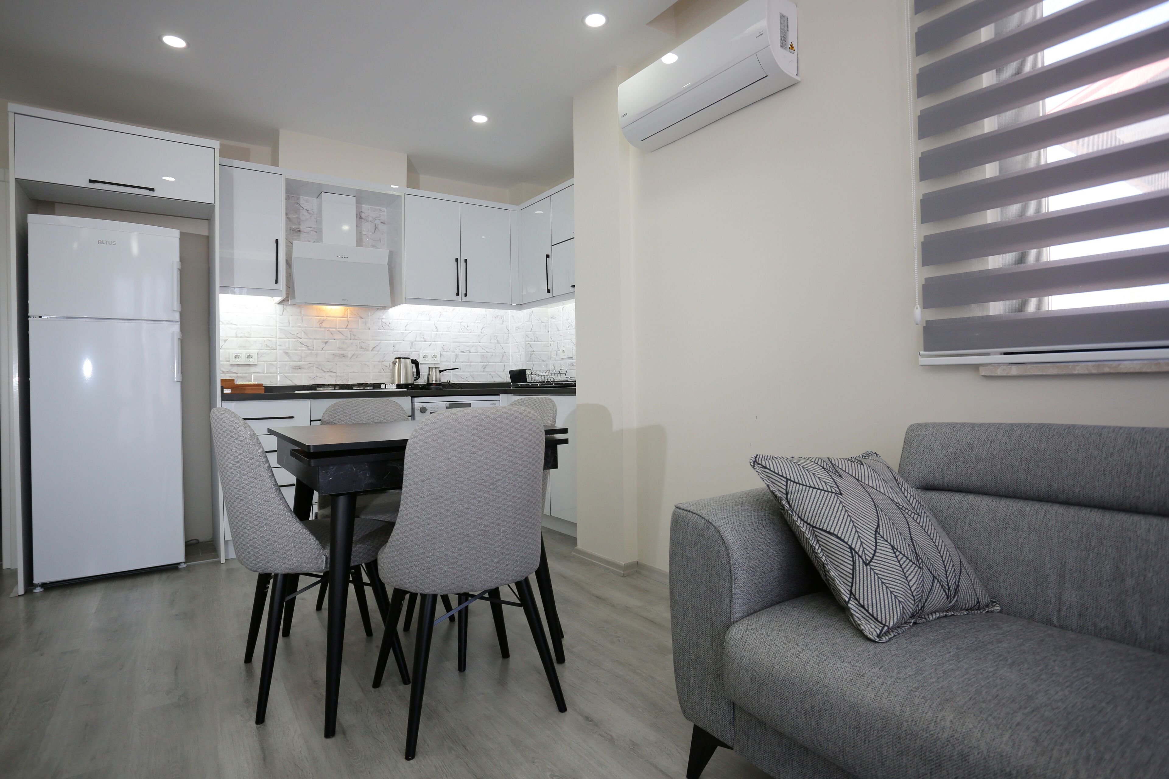 Une cuisine moderne avec des meubles blancs, une table à manger avec des chaises et un canapé gris agrémenté d’un coussin à motifs.