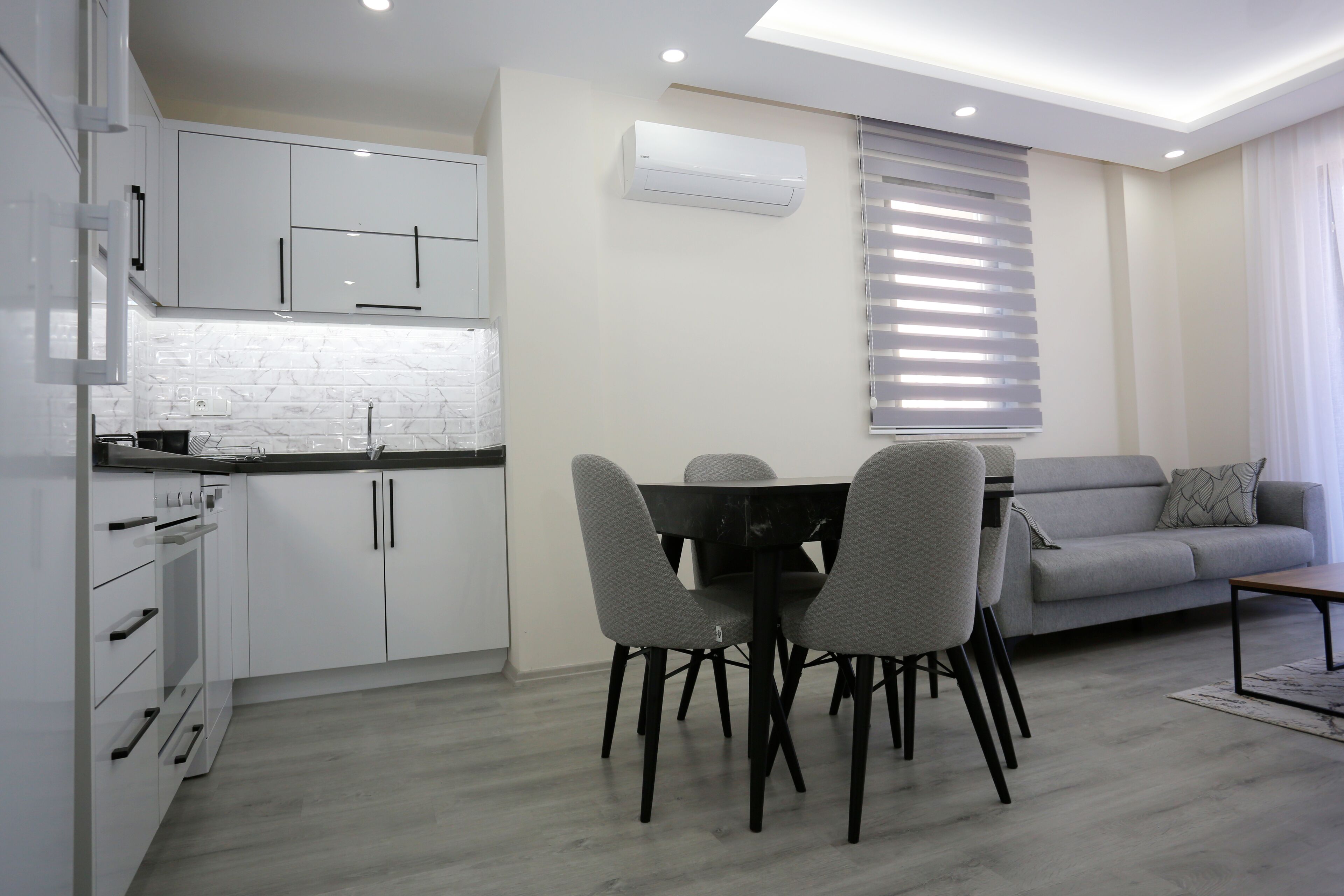 Een moderne keuken met witte kasten, een eettafel met stoelen en een grijze bank in een zithoek.