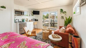 A modern, well-lit room with a kitchenette, a sofa, a dining table, and a view of a canal. - Bung-a-flo Tiny Bohemian Designed Floating Home w/ 360 Waterfront Views (Arnold)