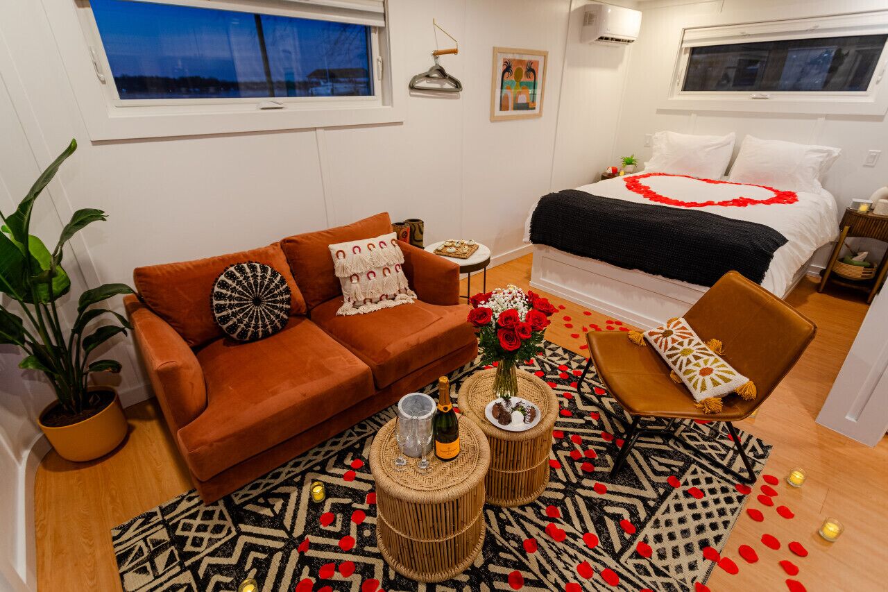 A cozy room with a brown sofa, a bed with a heart-shaped decoration, a small table with a vase of flowers, and a patterned rug.